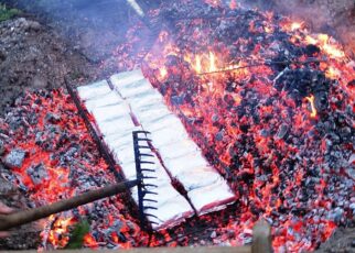 Absenken eines Drahtkorbes mit 20 Portionen Lakefleisch in die Glut. Das Fleisch ist in Alufolie eingewickelt und wird anschließend mit der Glut überschichtet.