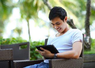 Mann sitzt auf einem Stuhl auf einer Holzterrasse unter Palmen und schaut auf ein Tablet. Vor in einem Tisch mit einem Softgetränk