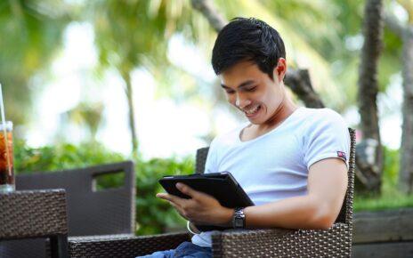 Mann sitzt auf einem Stuhl auf einer Holzterrasse unter Palmen und schaut auf ein Tablet. Vor in einem Tisch mit einem Softgetränk