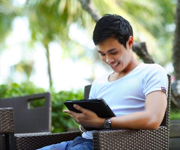 Mann sitzt auf einem Stuhl auf einer Holzterrasse unter Palmen und schaut auf ein Tablet. Vor in einem Tisch mit einem Softgetränk