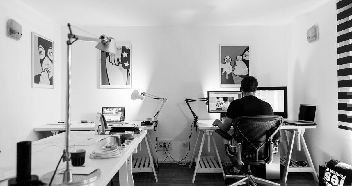 Blick ins Home-Office. Links ein Schreibtisch mit Lampe, hinten ein Mensch am Computer an einem weiteren Schreibtisch