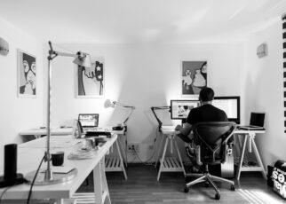 Blick ins Home-Office. Links ein Schreibtisch mit Lampe, hinten ein Mensch am Computer an einem weiteren Schreibtisch