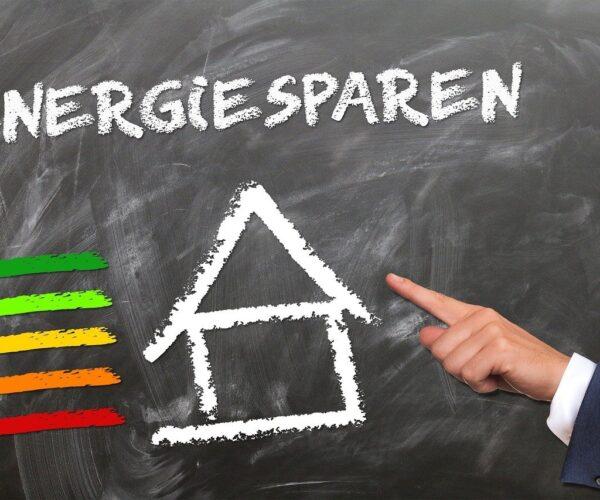 Energiesparen. Haus auf eine Tafel gemalt. Daneben die Farben der Energieeffizienzsymbole-Eine Hand mit ausgestrecktem Zeigefinder zeigt auf die Tafel