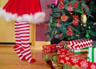 Festlich geschmückter Weihnachtsbaum mit einem Kind davor, welches aus Zehenspitzen steht. Vor dem Baum liegen bunt eingepackte Geschenke