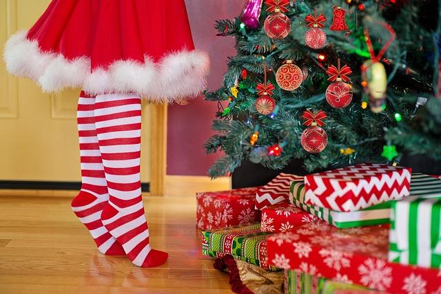 Festlich geschmückter Weihnachtsbaum mit einem Kind davor, welches aus Zehenspitzen steht. Vor dem Baum liegen bunt eingepackte Geschenke